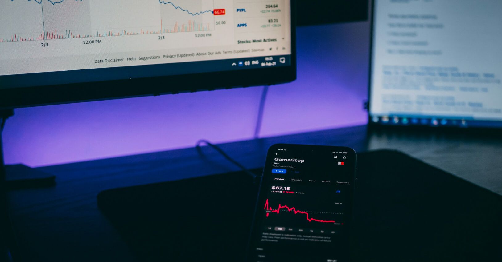 Smartfon z aplikacją giełdową wyświetlającą wykres akcji na tle monitora z danymi finansowymi i analizami rynkowymi.